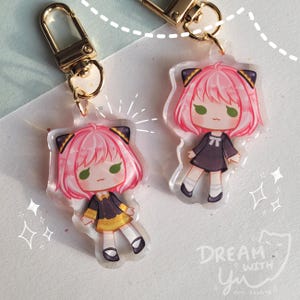 Peut inclure: Deux porte-clés en acrylique transparent représentant un personnage de dessin animé avec des cheveux roses, des yeux verts et une robe noire et dorée. Le personnage a des oreilles de chat et tient un petit objet blanc. Les porte-clés sont attachés à des anneaux en métal doré.