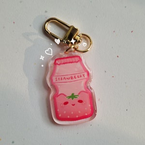 May include: A pink and white strawberry flavored yogurt keychain with a gold metal clasp. The keychain is made of clear acrylic and has a cartoon strawberry design.