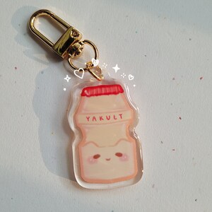 May include: A clear acrylic keychain with a cartoon illustration of a Yakult bottle. The bottle is white with a red lid and the word "Yakult" printed on the front. The keychain has a gold metal clasp.