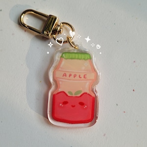 May include: A clear plastic keychain charm shaped like a small milk carton with a red apple design. The carton has a green lid and the word "APPLE" printed on the front. The charm has a gold metal ring attached to the top.