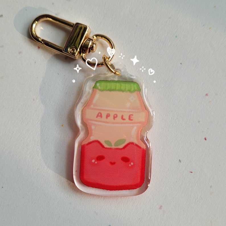 May include: A clear plastic keychain charm shaped like a small milk carton with a red apple design. The carton has a green lid and the word "APPLE" printed on the front. The charm has a gold metal ring attached to the top.