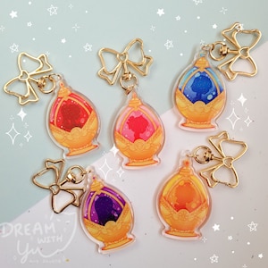 May include: Five colorful keychains featuring a gold-colored design with a clear glass orb containing a silhouette of a person. Each keychain has a gold-colored bow attached to a gold-colored ring.