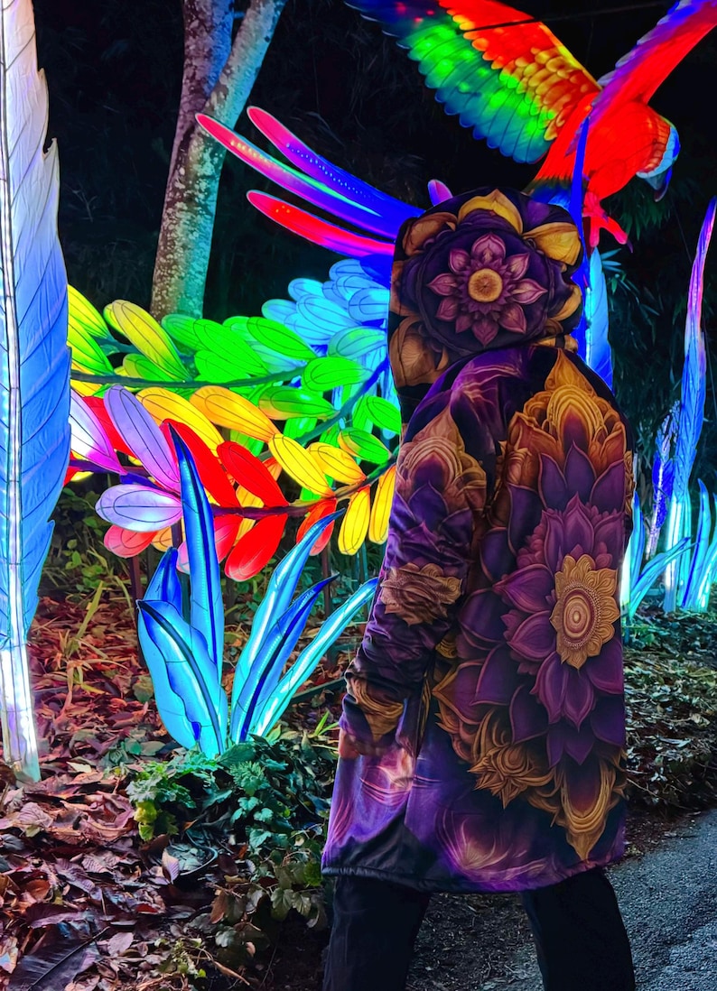 May include: A person wearing a purple and gold hooded jacket with a floral pattern. The jacket has a large flower design on the back. The person is standing in front of a colorful light display of flowers and a parrot.
