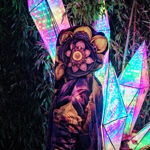 May include: A hooded jacket with a colorful floral print, featuring a large lotus flower design. The jacket is worn by a person standing in front of a background of iridescent, geometric light sculptures.