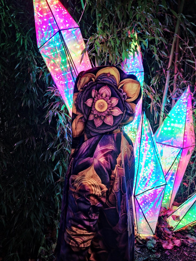 May include: A hooded jacket with a colorful floral print, featuring a large lotus flower design. The jacket is worn by a person standing in front of a background of iridescent, geometric light sculptures.