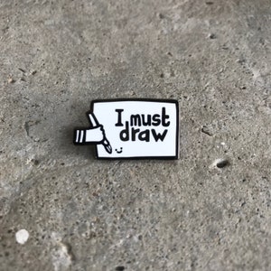 Pode incluir: Pin de esmalte preto e branco com uma mão segurando um lápis e desenhando um rosto sorridente em um retângulo branco com o texto "I must draw" nele.
