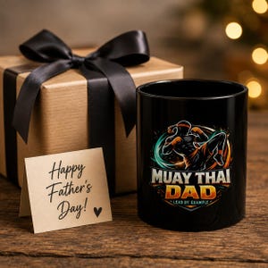 May include: A black ceramic mug featuring a graphic of a Muay Thai fighter in motion with the text "MUAY THAI DAD LEAD BY EXAMPLE" alongside a gift box with a black ribbon and a small card with the handwritten text "Happy Father's Day!" and a heart.