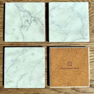 Op de afbeelding: Vier vierkante marmeren onderzetters met een wit en grijs geaderd patroon. Eén onderzetter toont een kurken achterkant met de tekst "the stonemason" en een logo. De onderzetters zijn gerangschikt op een houten oppervlak.