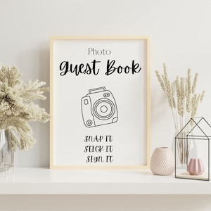 May include: A white sign with a black frame that says "Photo Guest Book" with a black and white illustration of a camera and the text "Snap It, Stick It, Sign It".