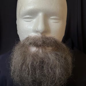 May include: A long, thick, brown beard made of synthetic hair. The beard is attached to a white mannequin head.