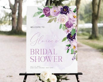Op de afbeelding: Een wit bord met paarse aquarelbloemen en tekst die luidt "Welkom bij de bruiloftsshower van Gloria op 17 juli 2027 Saratoga Springs, New York"