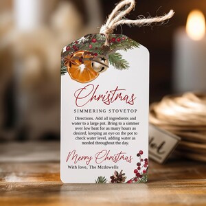 Op de afbeelding: Een wit, printbaar cadeau-etiket met een kerstthema. Het etiket heeft een rood en groen bloemmotief met een gedroogde sinaasappelschijf, kaneelstokjes en bellen. De tekst op het etiket luidt "Christmas Simmering Stovetop" en "Merry Christmas".