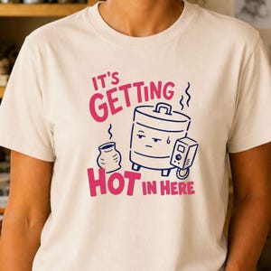Op de afbeelding: Een crèmekleurig t-shirt met een afbeelding van een dampende pot en een kleine verwarmer, inclusief de tekst "IT'S GETTING HOT IN HERE" in roze en donkerblauw. Het ontwerp toont een pot in cartoonstijl met een vermoeide gezichtsuitdrukking.