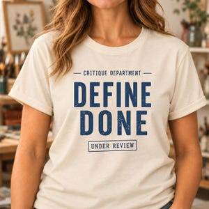 May include: A cream-colored short-sleeve t-shirt featuring dark blue text that reads CRITIQUE DEPARTMENT, DEFINE DONE, and UNDER REVIEW in a box. The design has a distressed, weathered texture on the lettering and graphics.