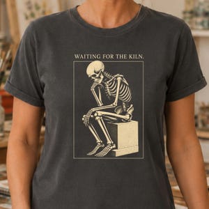May include: A charcoal gray short-sleeve t-shirt featuring a cream-colored graphic of a skeleton sitting on a block, resting its chin on its hand. Above the skeleton, the text reads "WAITING FOR THE KILN." in a simple, clean, capitalized font.
