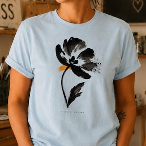 Puede incluir: Una camiseta de manga corta de color azul claro con un gráfico de flor hecho con pinceladas negras y un pequeño toque amarillo. El texto "STUDIO GROWN" está impreso en letras negras pequeñas debajo del diseño floral. Tiene cuello redondo.