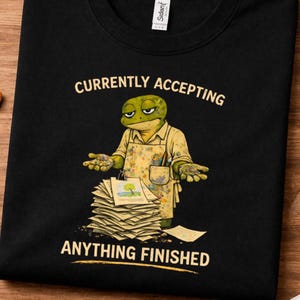 May include: Black t-shirt featuring a cartoon frog in an artist's apron. The text says "CURRENTLY ACCEPTING ANYTHING FINISHED". The frog is positioned in front of a pile of papers, with a small painting on top.