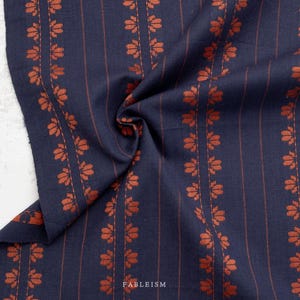 May include: A dark navy blue fabric featuring vertical stripes and repeating rows of small, rust-colored floral motifs. The textile displays a woven texture with the word FABLEISM printed in small white letters near the center of the fabric.