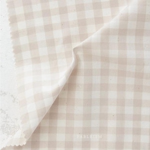 May include: A piece of beige and white gingham fabric with a raw edge. The fabric is folded over, revealing the pattern and texture. The word "FABLEISM" is printed in the bottom right corner.
