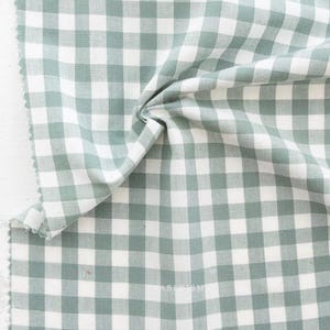 May include: A close-up of a piece of fabric with a gingham pattern in white and a muted green color. The fabric is folded and draped, showing the texture and weave of the material. The edges are unfinished.