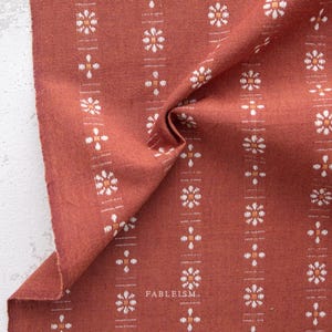 May include: Close-up of a folded terracotta-colored fabric with a repeating pattern of white and orange floral designs. The fabric has a textured appearance and the word "FABLEISM" is printed on the fabric.