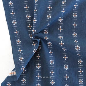 May include: Close-up of a navy blue fabric with a repeating floral pattern. The design features white and orange flowers arranged in vertical stripes. The fabric is folded, showing texture and detail. The word "FABLEISM" is printed on the fabric.