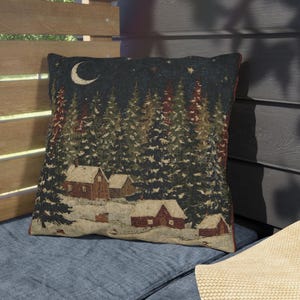 May include: A square throw pillow featuring a woven winter scene of snow-covered cabins nestled among dark green pine trees under a night sky with a crescent moon and stars. The design displays a muted color palette of deep green, brown, and white.