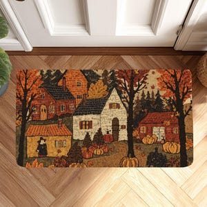 May include: A rectangular doormat featuring a printed autumn scene with three houses, tall trees with orange and brown foliage, and several orange pumpkins scattered across the ground. The illustration includes a small figure standing near a yellow house.