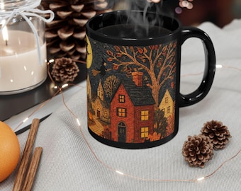 May include: A black ceramic mug featuring a detailed autumnal scene with a red house, a yellow house, a church, and a witch flying on a broomstick against a full moon. The design includes bare trees and orange foliage, creating a festive autumn atmosphere.