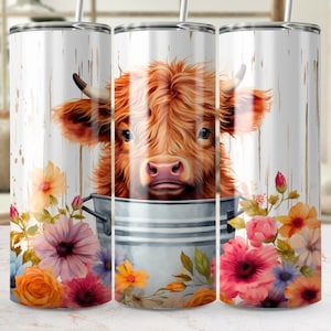 May include: A white stainless steel tumbler with a brown and white Highland cow peeking out of a metal bucket. The cow is surrounded by colorful flowers, including orange, pink, purple, and yellow.