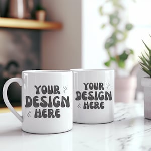 Op de afbeelding: Twee witte keramische mokken met zwarte tekst die "Your Design Here" zegt op een wit marmeren oppervlak.