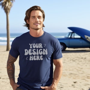 May include: A man wearing a navy blue t-shirt with the text "YOUR DESIGN HERE" in a retro font. He is standing on a sandy beach with a blue car and surfboard in the background.