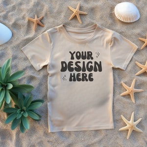 May include: A beige t-shirt with the text "YOUR DESIGN HERE" in a retro font. The shirt is lying on a sandy surface with seashells and starfish.