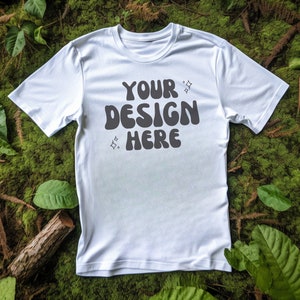 May include: White t-shirt with the text "YOUR DESIGN HERE" in a retro font on a green mossy background.