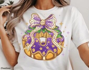 May include: White t-shirt with a purple, green, and gold donut with a gold bow on top. The donut has gold stars and polka dots.