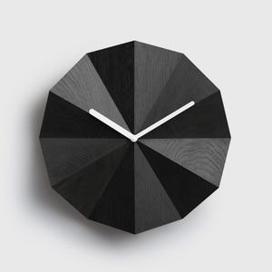 Może przedstawiać: Dwunastokątny zegar ścienny z czarnym wykończeniem o strukturze drewna i fasetowaną, geometryczną powierzchnią. Tarcza zegara zawiera dwie cienkie, białe wskazówki umieszczone centralnie, kontrastujące z ciemnymi, teksturowanymi panelami drewnianymi.