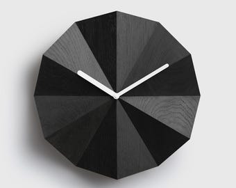 Puede incluir: Un reloj de pared dodecagonal con acabado de veta de madera negra y una superficie geométrica facetada. La esfera del reloj incluye dos manecillas blancas delgadas centradas en el medio, que contrastan con los paneles de madera oscuros y texturizados.