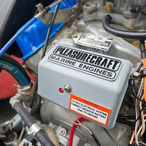 May include: Close-up of a gray electrical box with the text "PLEASURECRAFT MARINE ENGINES" on the front. A red warning label with the text "CAUTION 12 VOLT ELECTRICAL SYSTEM CONNECT NEGATIVE BATTERY LEAD TO GROUND" is attached to the box.