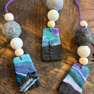 May include: Three handmade car air fresheners with a purple, blue, and green aurora borealis design. Each air freshener is attached to a string with wooden beads and gray felt balls.