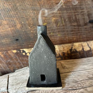 May include: A small, black ceramic house-shaped incense burner with a square base. Smoke rises from the chimney. The house has a small rectangular opening and a textured surface. The background is a wooden surface.