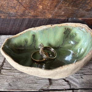 May include: A green ceramic ring dish with a rough, textured exterior. Two gold rings are resting in the dish.