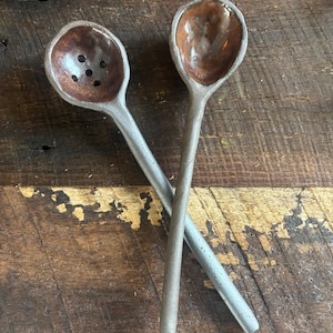 May include: Two ceramic spoons with brown glaze. One spoon has holes in the bowl. The spoons are on a wooden surface.
