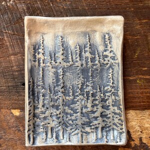 May include: A rectangular ceramic dish with a forest scene. The dish features a raised relief of pine trees in shades of gray and blue. The edges are a light beige color, and the dish is set on a wooden surface.