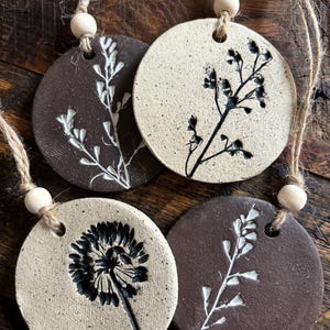 May include: Four round ceramic ornaments with a rustic, speckled finish. Each ornament features a different pressed floral design in black or white. The ornaments are strung with twine and wooden beads.