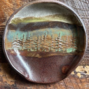 May include: A round ceramic spoon rest featuring a textured forest scene with embossed pine trees. The glaze transitions from a deep dark brown at the base to a vibrant teal and green middle section, topped with a creamy off-white rim.