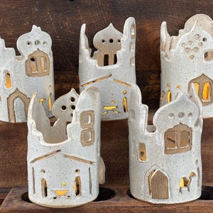May include: Five ceramic candle holders in a light gray color, shaped like Middle Eastern buildings. Each has cut-out windows and doors, with a warm glow emanating from within. The holders are arranged on a wooden surface.