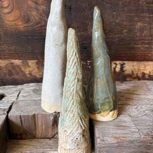 May include: Three ceramic cone-shaped sculptures in varying sizes and colors. The tallest is off-white with speckles, the middle one is textured with a light blue and tan pattern, and the third is teal with gold accents. All have a natural, rustic aesthetic.