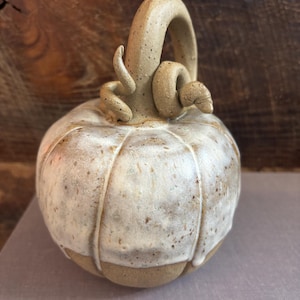 May include: A ceramic pumpkin sculpture featuring a speckled tan clay body with a white glazed top section that drips down the sides. The pumpkin has a curved, textured stem with a small, coiled vine detail attached at the base of the stem.