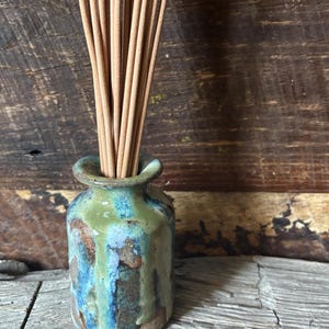May include: A small, cylindrical ceramic vase with a flared rim, featuring a reactive glaze in shades of sage green, deep blue, and earthy brown. A bundle of thin, light tan wooden incense sticks is placed upright inside the narrow opening.