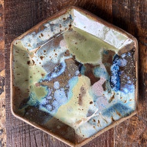 May include: A hexagonal ceramic dish featuring a glossy glaze with a mottled pattern of sage green, deep blue, brown, and white. The surface displays varied textures and speckles, with a raised rim and a smooth, reflective finish throughout.